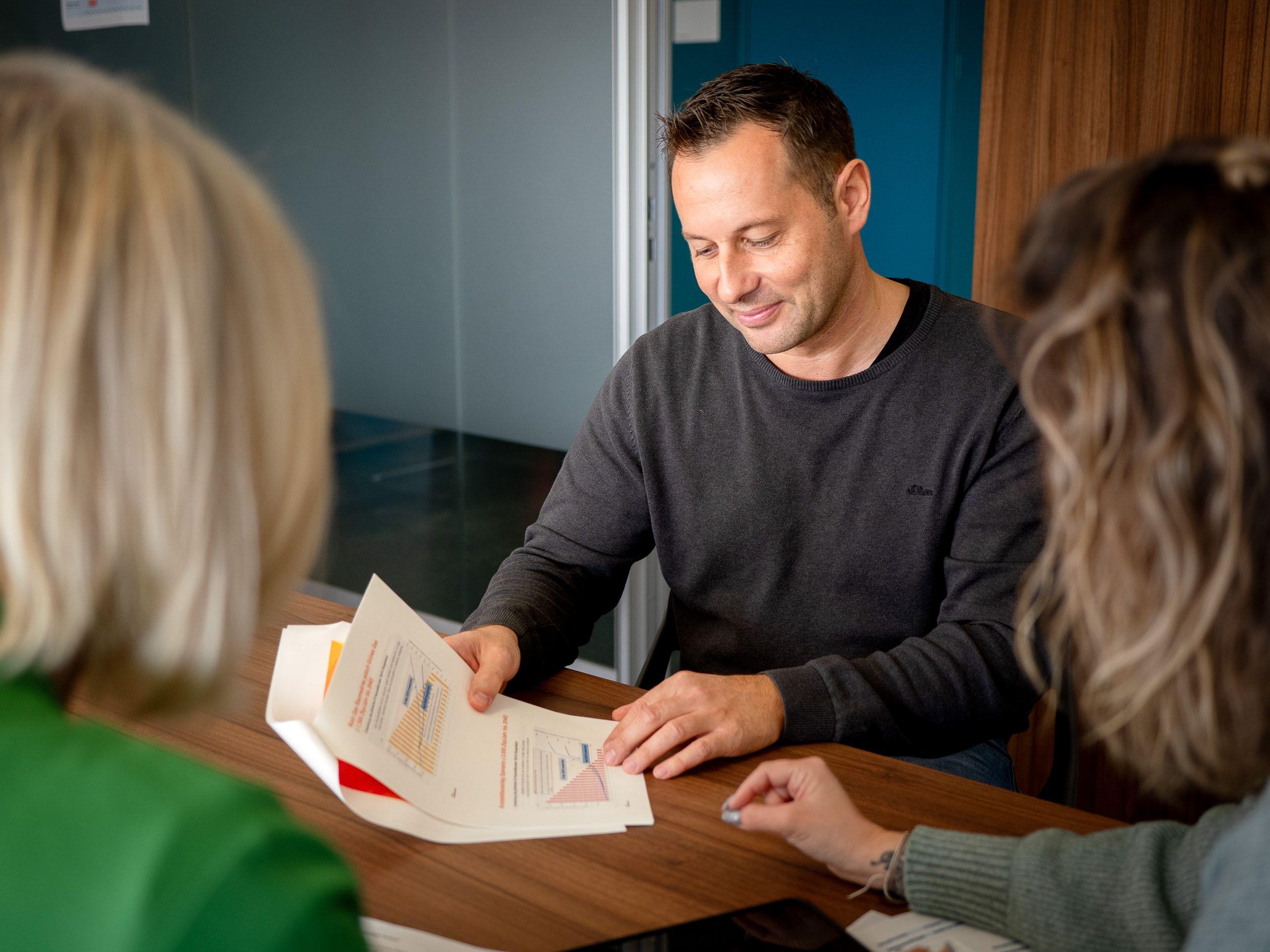 3 Personen sitzen am Tisch. Eine Person erklärt das Lastprofil des Stromverbrauchs anhand eines ausgedruckten Diagramms.