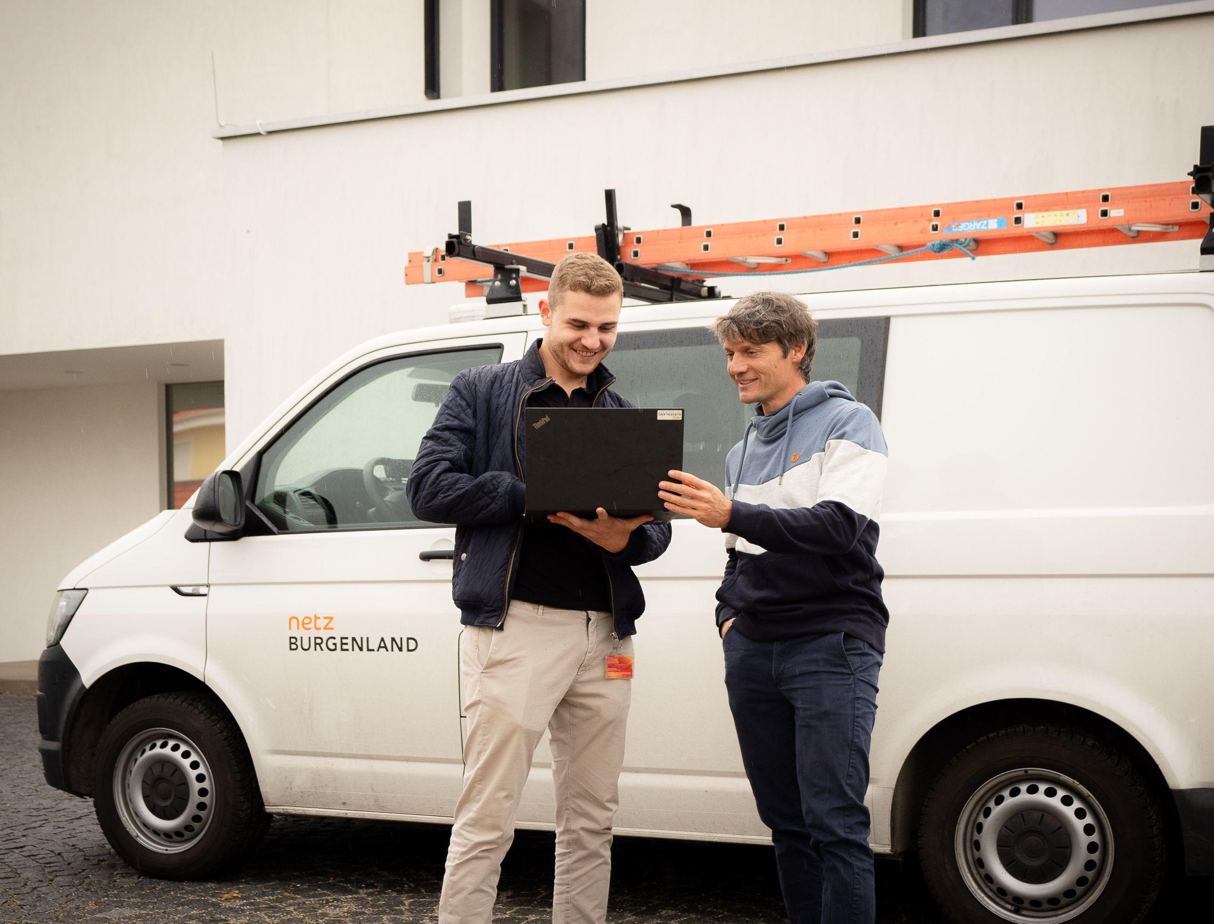 Zwei Personen mit einem Laptop besprechen etwas neben einem Dienstwagen mit der Aufschrift Netz Burgenland