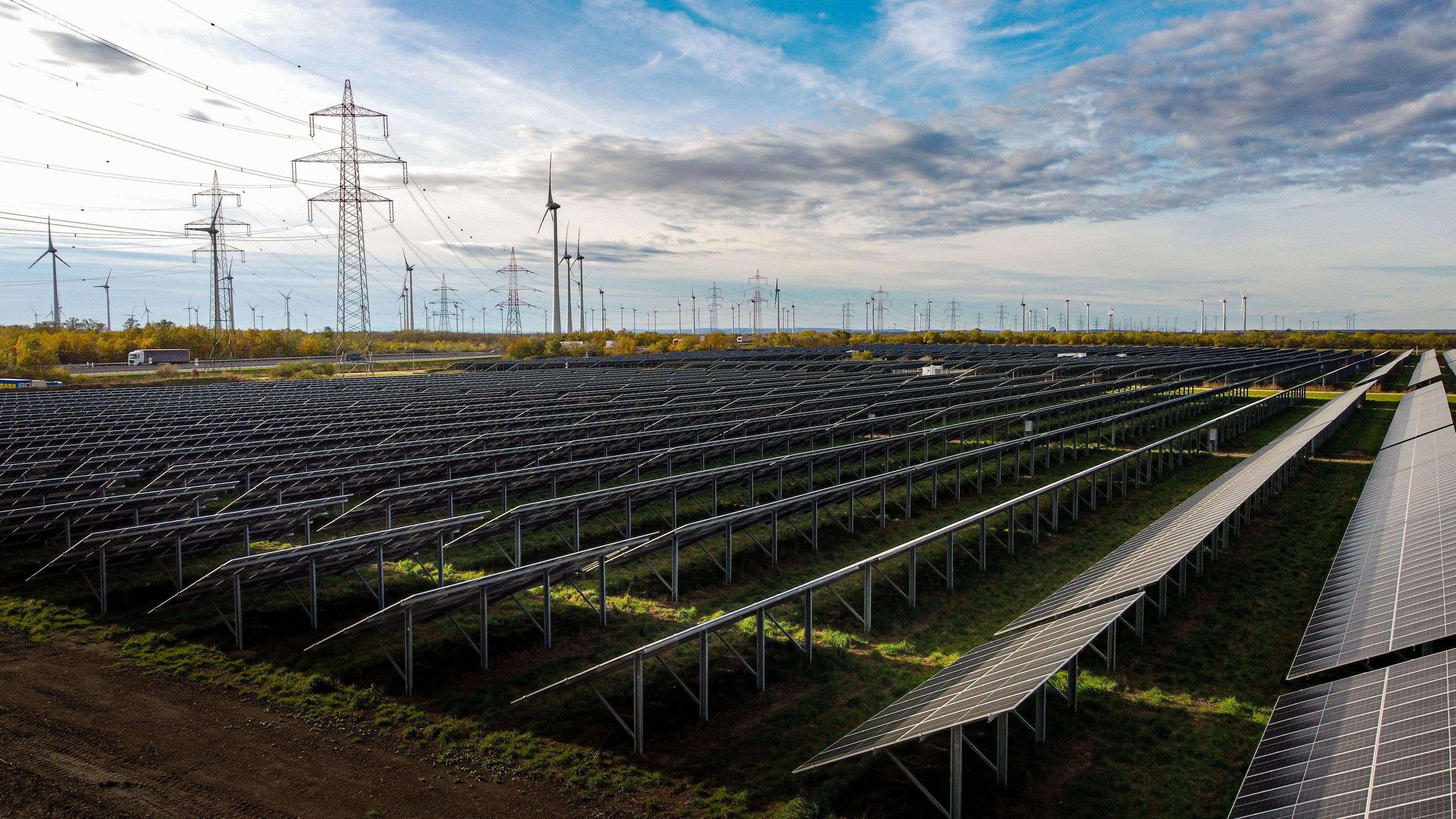 Photovoltaikmodule im Vordergrund mit Windkraftanlagen und Hochspannungsleitungen im Hintergrund vor einem bewölkten Himmel.