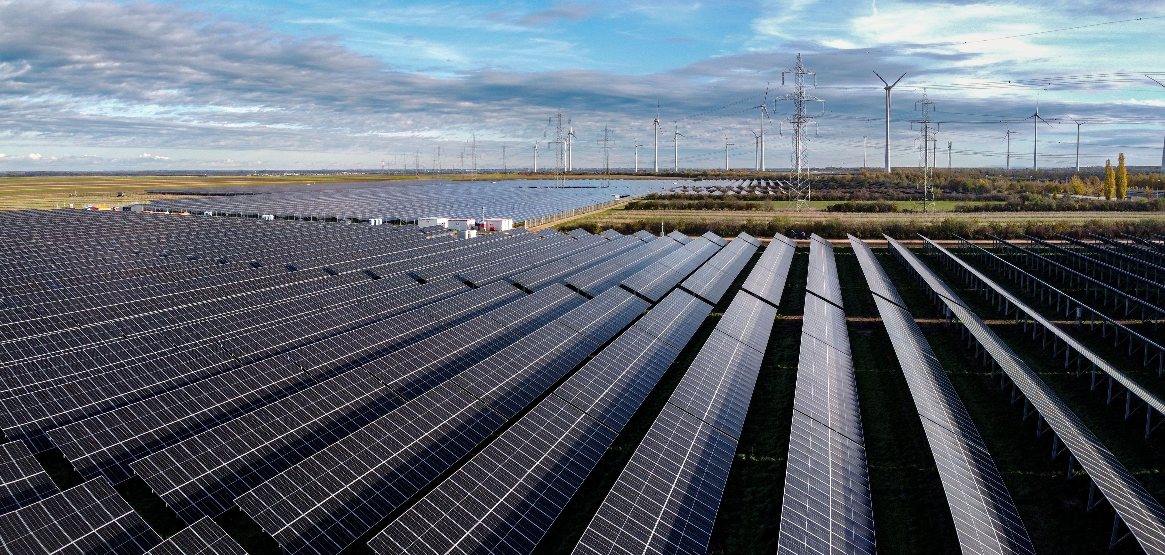 Luftaufnahme eines großen Solarparks mit Reihen von Solarmodulen und Windrädern im Hintergrund