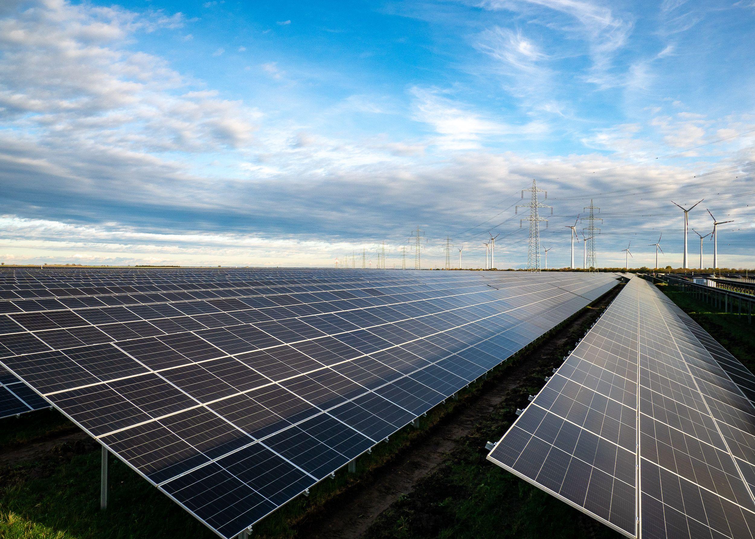 Endlose Reihen von Solarpaneelen erstrecken sich bis zum Horizont, begleitet von Windkraftanlagen und Hochspannungsmasten unter einem dynamischen Himmel.