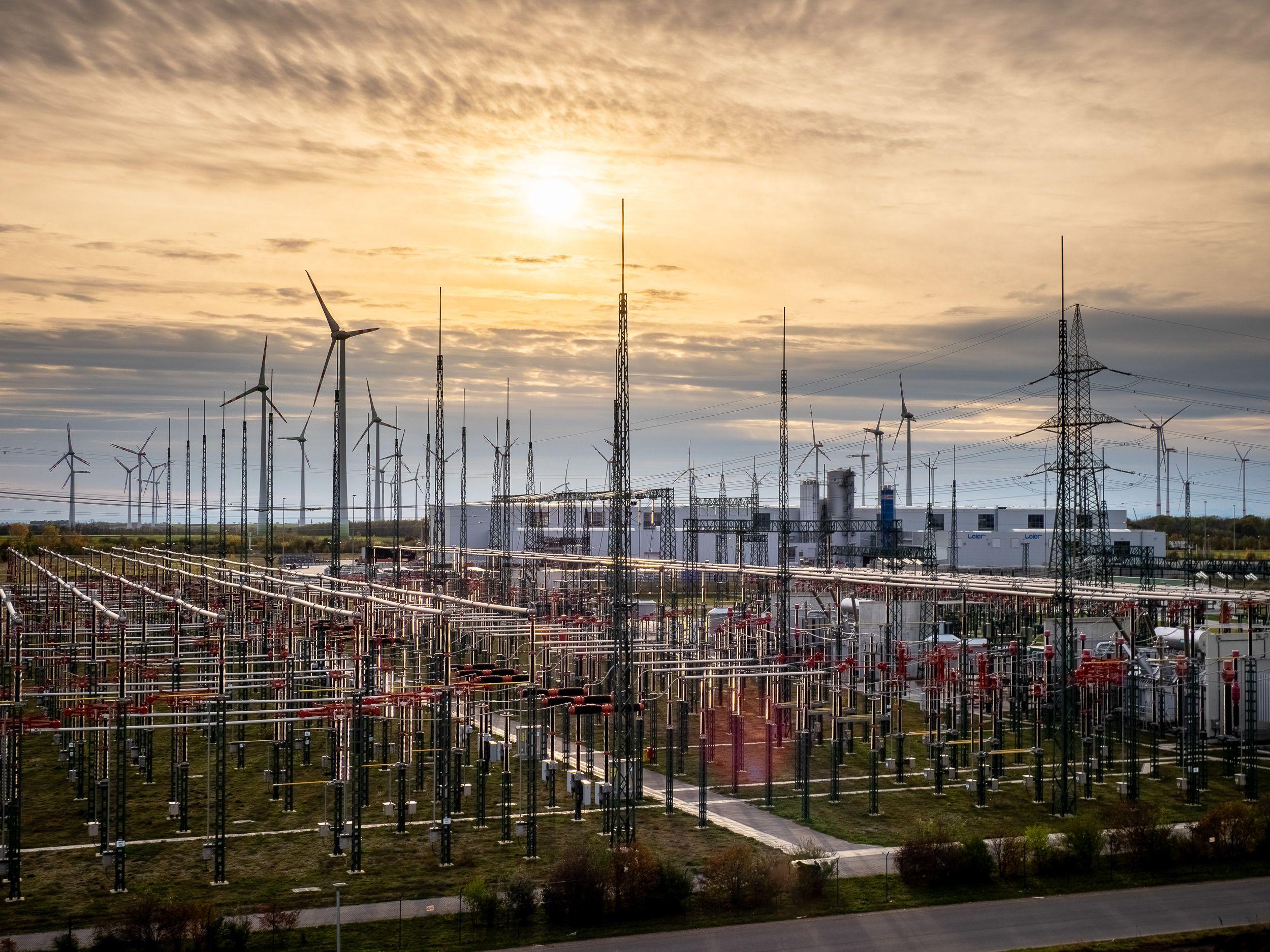 Umspannwerk mit Windrädern und Hochspannungsmasten im Gegenlicht eines malerischen Sonnenuntergangs.