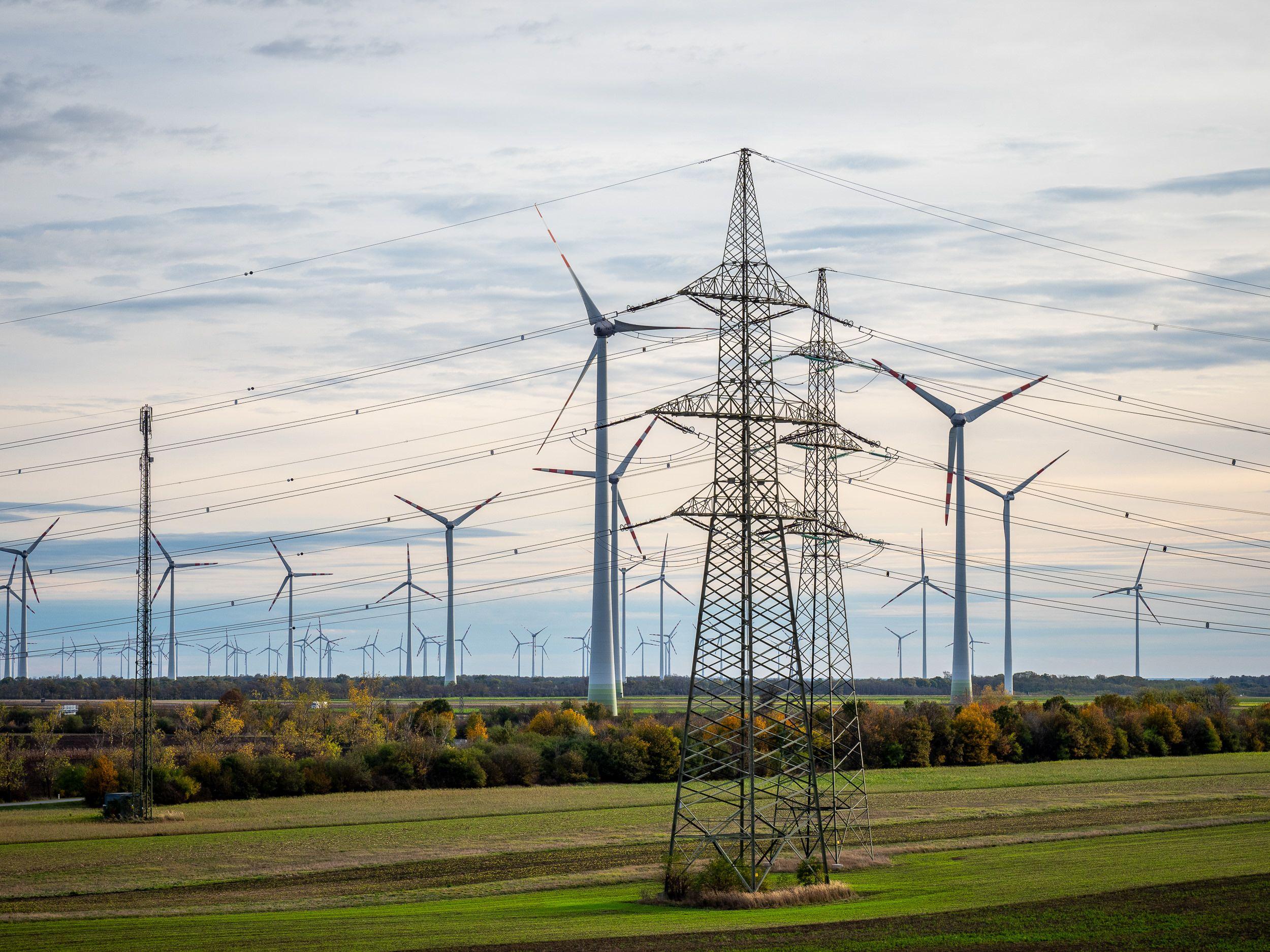 Ein elektrischer Hochspannungsmast vor einem Feld mit Windkraftanlagen im Hintergrund.