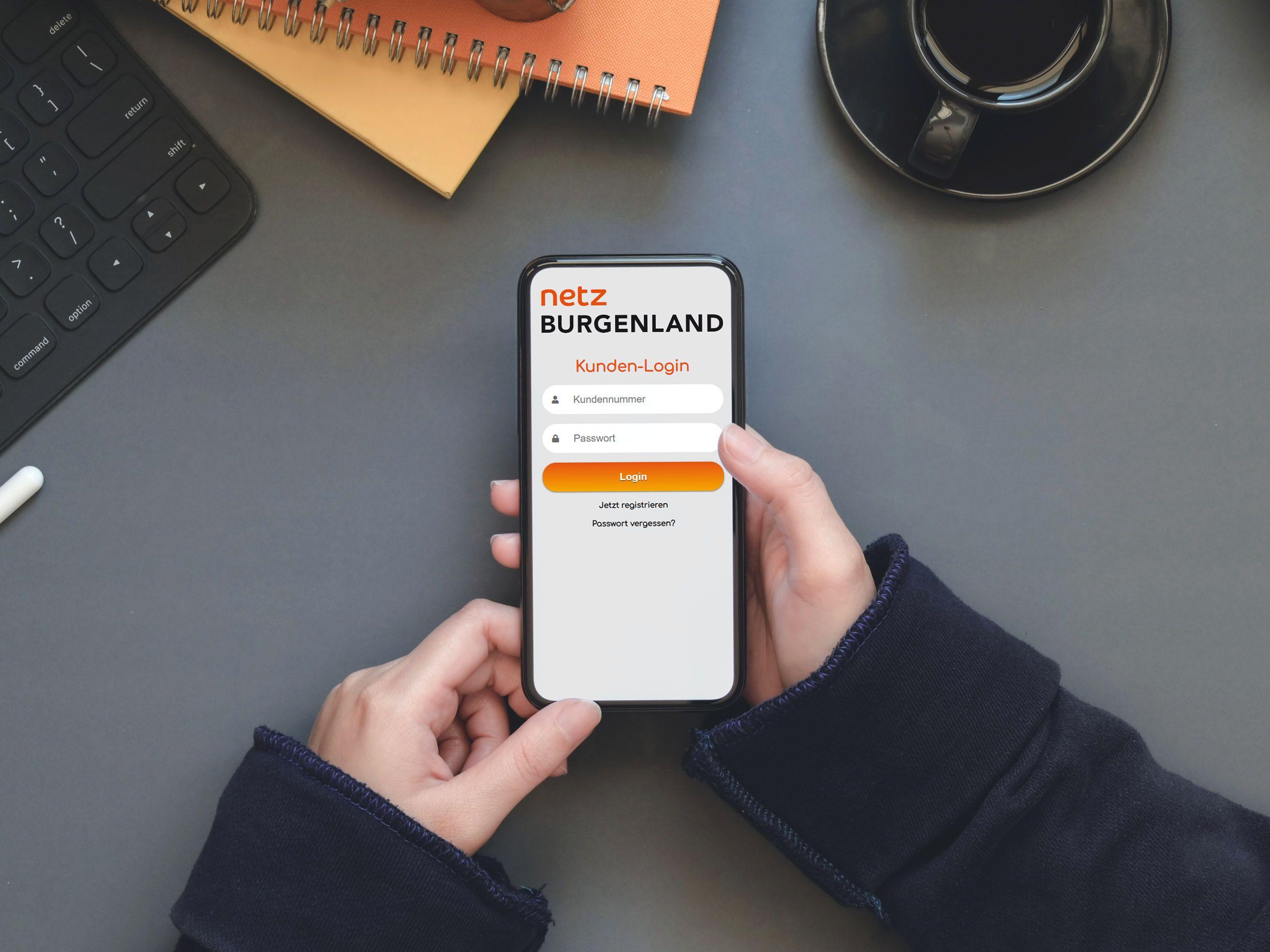 Smartphone mit geöffnetem Kundencenter-Login der Netz Burgenland auf dem Bildschirm, neben Tastatur und Kaffeebecher auf einem Schreibtisch.